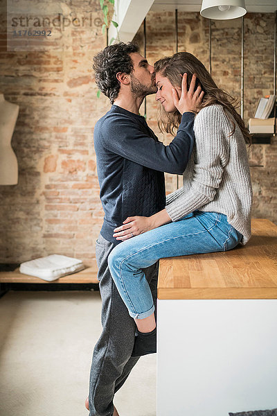 Junger Mann küsst Freundin auf der Küchenbank auf die Stirn