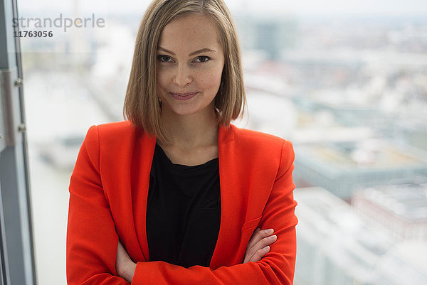 Porträt einer jungen Geschäftsfrau mit verschränkten Armen vor einem Bürofenster