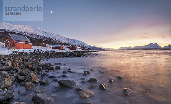 Panorama der typischen Holzhütten namens Rorbu  eingerahmt von feurigem Himmel bei Sonnenuntergang und gefrorenem Meer  Djupvik  Lyngen Alps  Troms  Norwegen  Skandinavien  Europa