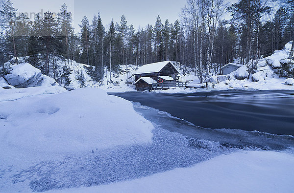 Die Dämmerung umrahmt das gefrorene Wasser im verschneiten Wald und eine Holzhütte  Juuma  Myllykoski  Region Lappland  Finnland  Europa