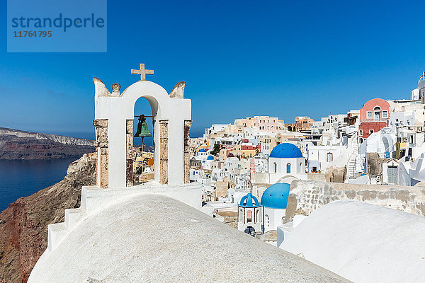 Ein Kirchendach und eine Glocke mit den weiß getünchten Steinmauern und blauen Kirchenkuppeln von Oia  Santorin  Kykladen  Griechische Inseln  Griechenland  Europa
