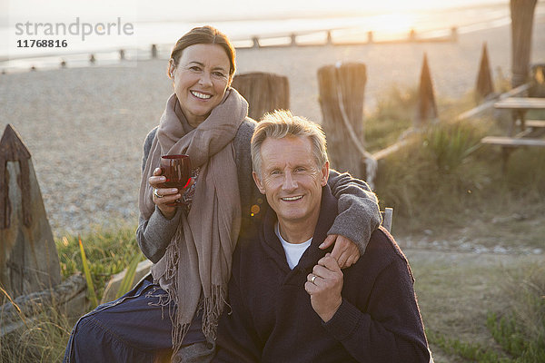 Portrait lächelndes reifes Paar mit Händchenhalten und Weintrinken am Strand bei Sonnenuntergang