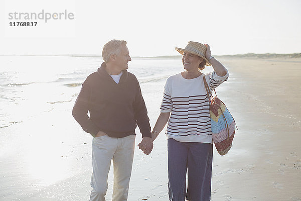 Lächelndes reifes Paar hält sich an den Händen und geht am sonnigen Strand spazieren.