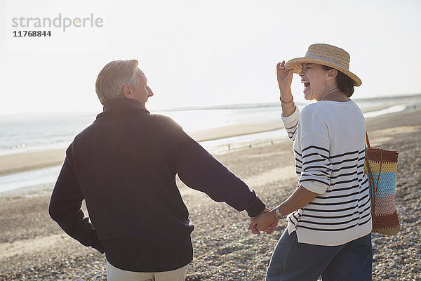 Lachendes reifes Paar  das Händchen hält und am sonnigen Strand spazieren geht.