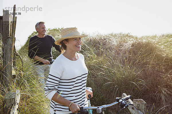 Ein reifes Paar geht mit dem Fahrrad am sonnigen Strand entlang.