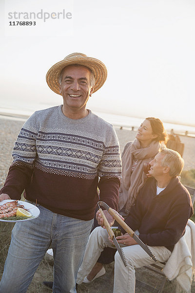 Portrait lächelnder älterer Mann beim Grillen mit Freunden am Strand bei Sonnenuntergang