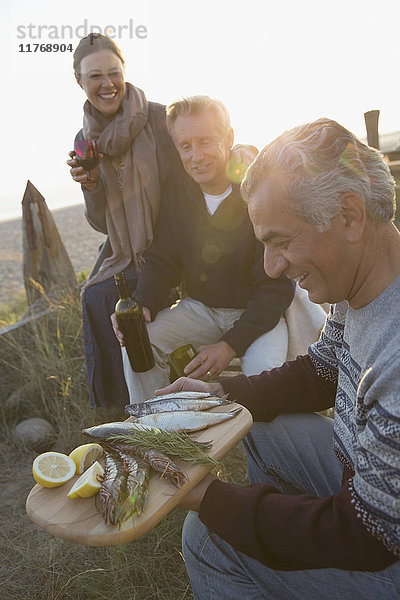 Reife Freunde trinken Wein und grillen Fische am Strand bei Sonnenuntergang.
