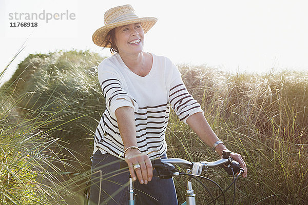 Lächelnde reife Frau beim Radfahren auf dem sonnigen Strandgrasweg
