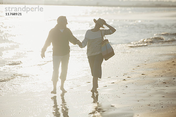 Ein reifes Paar hält sich an den Händen und geht am sonnigen Strand spazieren.
