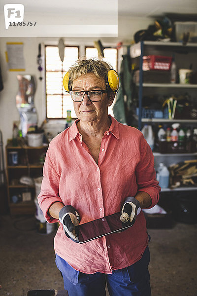 Nachdenkliche Seniorin mit digitalem Tablett im Stehen in der Werkstatt