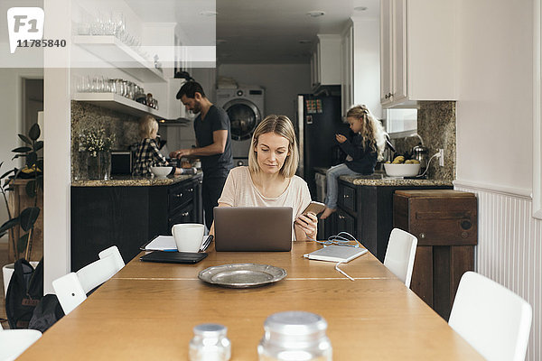 Mittlere erwachsene Frau  die das Handy hält  während sie den Laptop am Tisch mit der Familie in der Küche zu Hause benutzt.