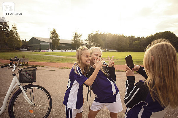 Mädchen beim Fotografieren von Freunden  die Handzeichen auf dem Fußweg am Fußballfeld zeigen.