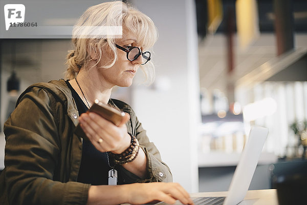 Geschäftsfrau  die ein Smartphone hält  während sie den Laptop am Schreibtisch im Büro benutzt.