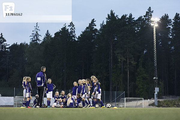 Trainer steht mit Mädchen-Fußballmannschaft auf dem Feld gegen Bäume