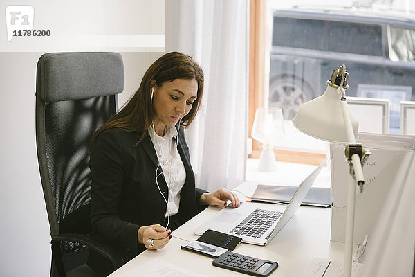 Reife Immobilienmaklerin hört Kopfhörer  während sie sich den Laptop im Büro ansieht.