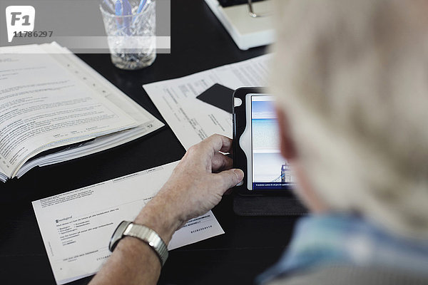 Abgeschnittenes Bild eines älteren Mannes  der beim Lesen von Rechnungen am Tisch ein digitales Tablett benutzt.