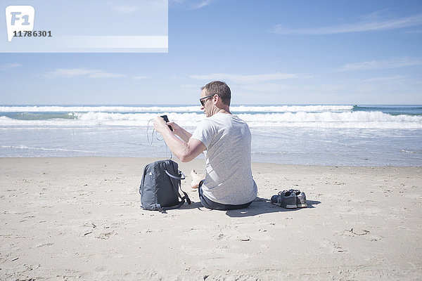 Rückansicht des Mannes mit Handy beim Herausnehmen der Kopfhörer aus dem Rucksack am Strand