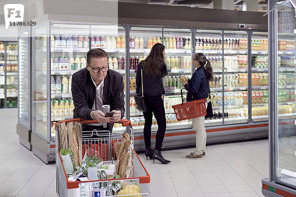 Reifer Mann  der das Handy benutzt  während er sich auf den Einkaufswagen lehnt  gegen Frauen  die Säfte im Kühlregal im Supermarkt kaufen.