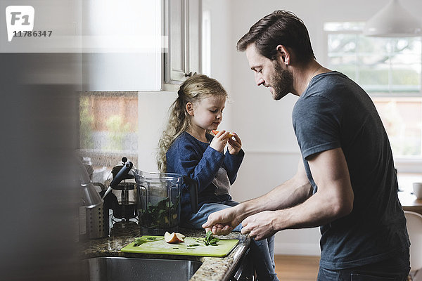 Seitenansicht des Vaters beim Kochen  während die Tochter einen Apfel in der Küche hat.