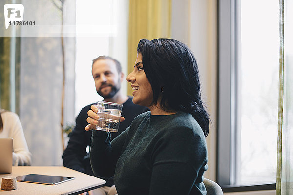 Lächelnde Geschäftsfrau hält ein Glas Wasser  während sie mit Kollegen am Konferenztisch sitzt.