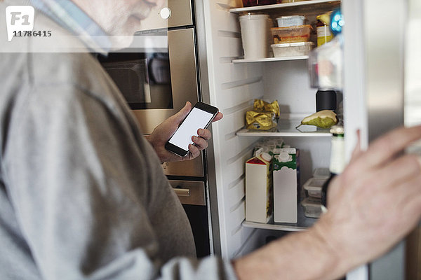 Senior Mann hält Telefon während der Suche im Kühlschrank zu Hause
