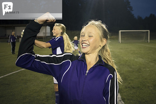 Porträt eines fröhlichen Mädchens  das seine Muskeln gegen Freunde auf dem Fußballfeld spannt.