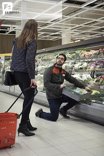 Verkäuferin kniend mit Kiste im Gespräch mit einer Frau  die im Supermarkt einen Korb hält