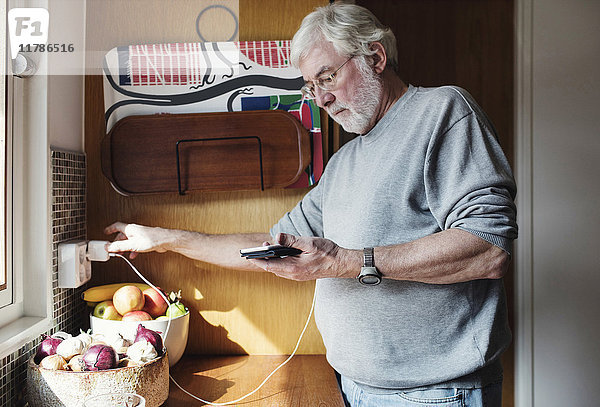 Senior Mann hält Handy  während er das Ladegerät an die Steckdose in der heimischen Küche anschließt.
