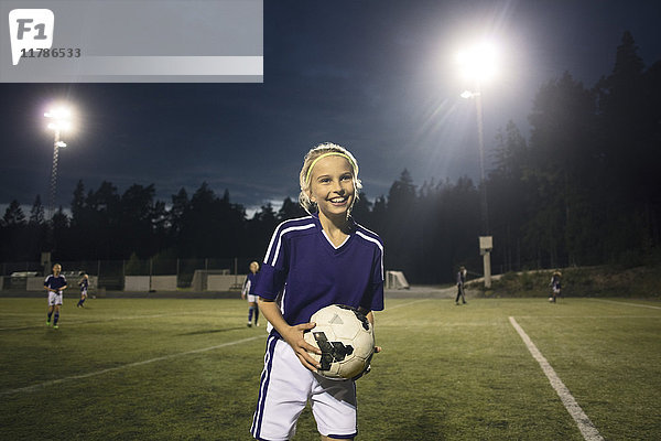 Glücklich  dass das Mädchen nachts mit dem Fußball auf dem Spielfeld gegen den Himmel steht.