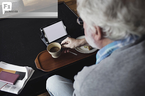 Abgeschnittenes Bild eines älteren Mannes  der eine Teetasse mit einem digitalen Tablett am Tisch hält.