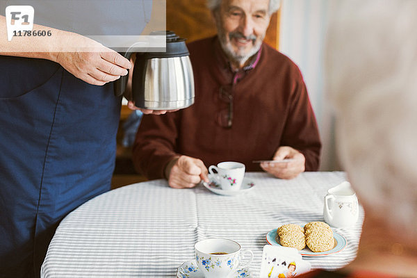 Mittelteil des Hausmeisters  der die Kaffeekanne hält  während das ältere Paar im Pflegeheim Karten spielt.