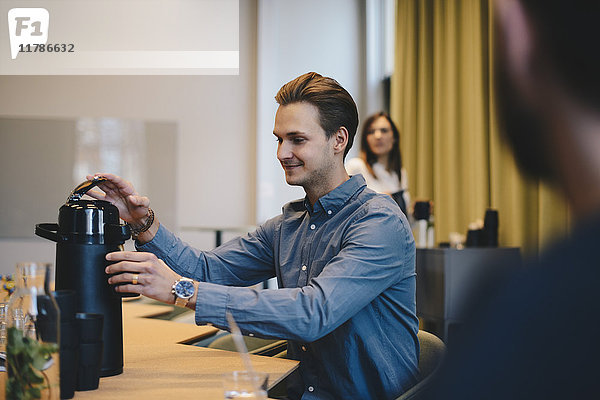Lächelnder Geschäftsmann öffnet isolierten Getränkebehälter am Konferenztisch mit Kollegen im Sitzungssaal