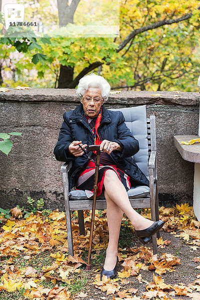 Volle Länge der älteren Frau  die auf einem Stuhl im Park sitzt und einen Stock hält.