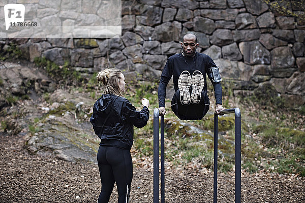 Rückansicht einer Sportlerin  die den Mann beim Training an parallelen Stäben gegen die Steinmauer im Wald beobachtet.