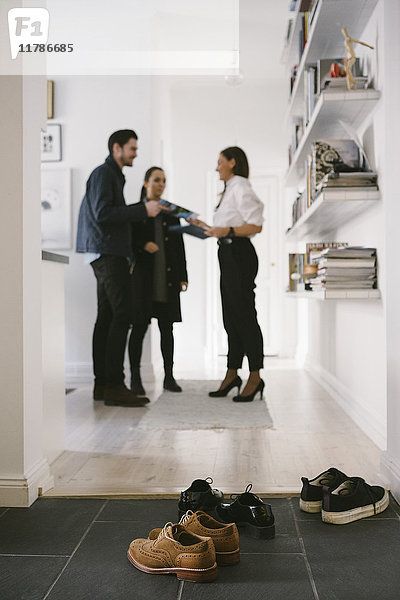 Verschiedene Schuhe auf dem Boden mit einer Maklerin  die mit einem jungen Paar über Regale im Hintergrund diskutiert.