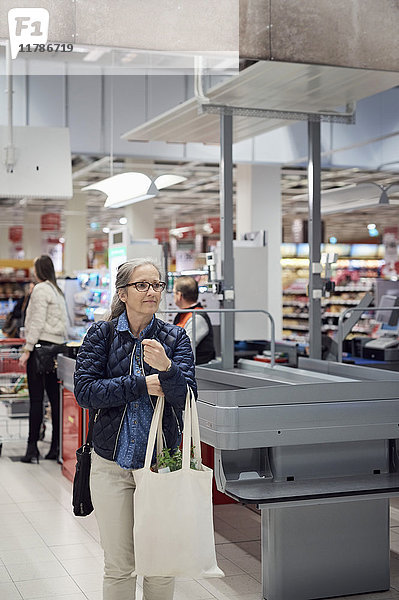 Reife Frau mit Einkaufstasche beim Gehen an der Kasse im Supermarkt
