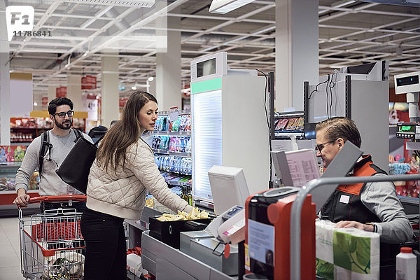 Mann schaut Frau an  die im Supermarkt für eine reife Kassiererin bezahlt.