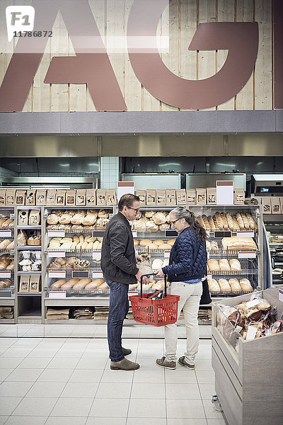 Erwachsenes Paar steht mit Korb gegen Brotgestell im Supermarkt