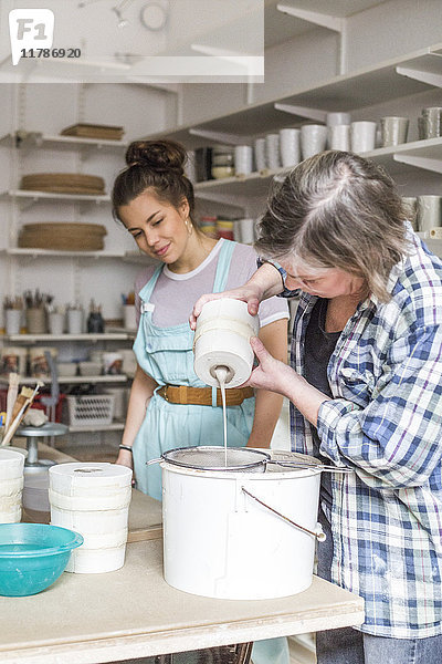 Junge Mitarbeiterin beim Anblick einer reifen Töpferin  die in der Werkstatt Ton aus einer Vase in einem Sieb auf einem Eimer gießt.