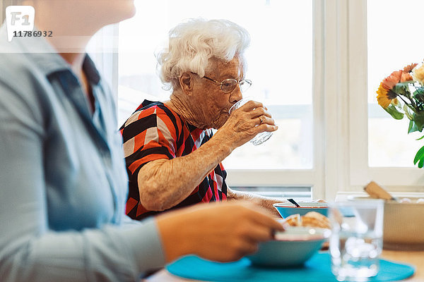 Seniorin trinkt Wasser  während sie mit ihrer Tochter am Esstisch sitzt.