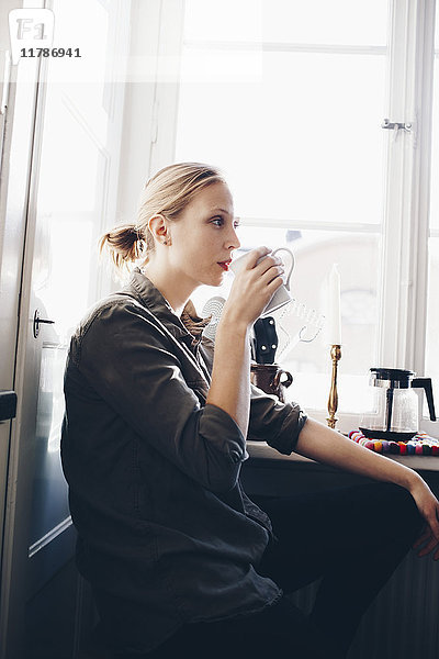 Seitenansicht der Frau beim Kaffeetrinken gegen das Fenster in der heimischen Küche