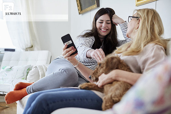 Mutter zeigt Handy  während die Tochter zu Hause mit dem Hund auf dem Sofa spielt.
