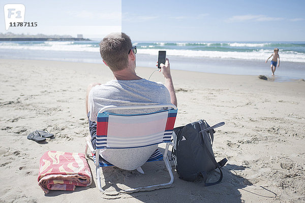 Rückansicht des Mannes auf dem Stuhl am Strand mit dem Handy