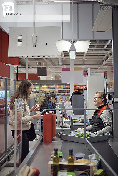 Frau im Gespräch mit reifer Kassiererin beim Bezahlen an der Kasse im Supermarkt