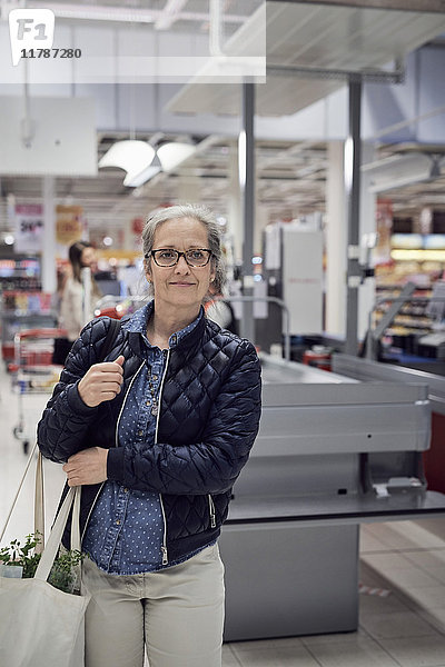 Lächelnde reife Frau mit Einkaufstasche beim Blick gegen die Kasse im Supermarkt