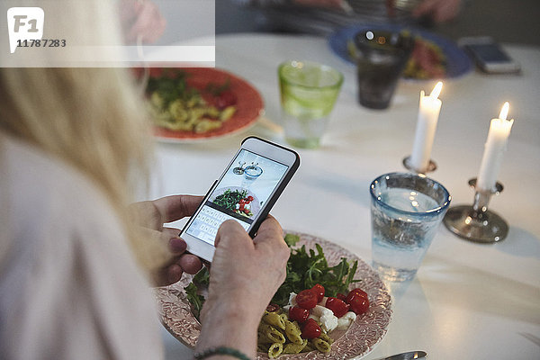 Abgeschnittenes Bild einer Frau  die Essen durch ein Smartphone am Esstisch fotografiert.