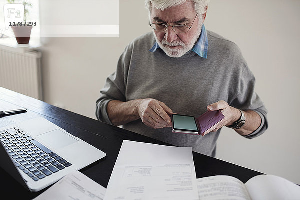 Senior Mann mit Laptop hält Handy beim Lesen von Rechnungen am Tisch