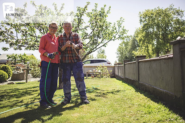 Ganzkörperporträt eines älteren Paares mit Hammer und Gartenschlauch im Hof stehend
