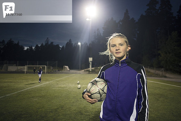 Porträt eines Mädchens mit Fußball auf dem Spielfeld gegen Bäume bei Nacht