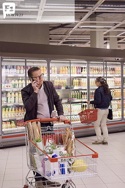 Der reife Mann spricht auf dem Handy  während die Frau auf die Vitrine im Kühlregal des Supermarktes schaut.